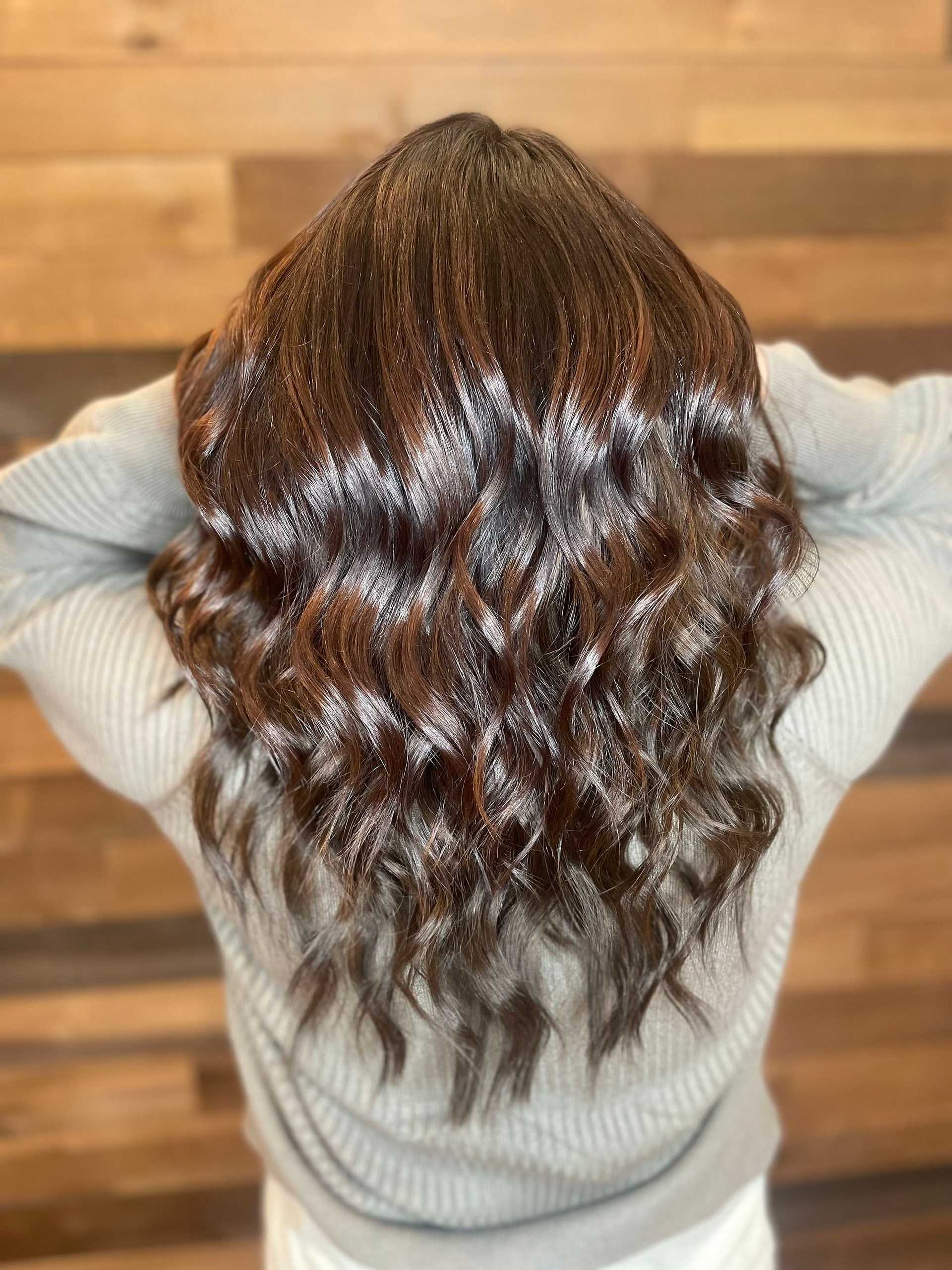 Woman with wavy, highlighted hair facing away, against a wooden background.